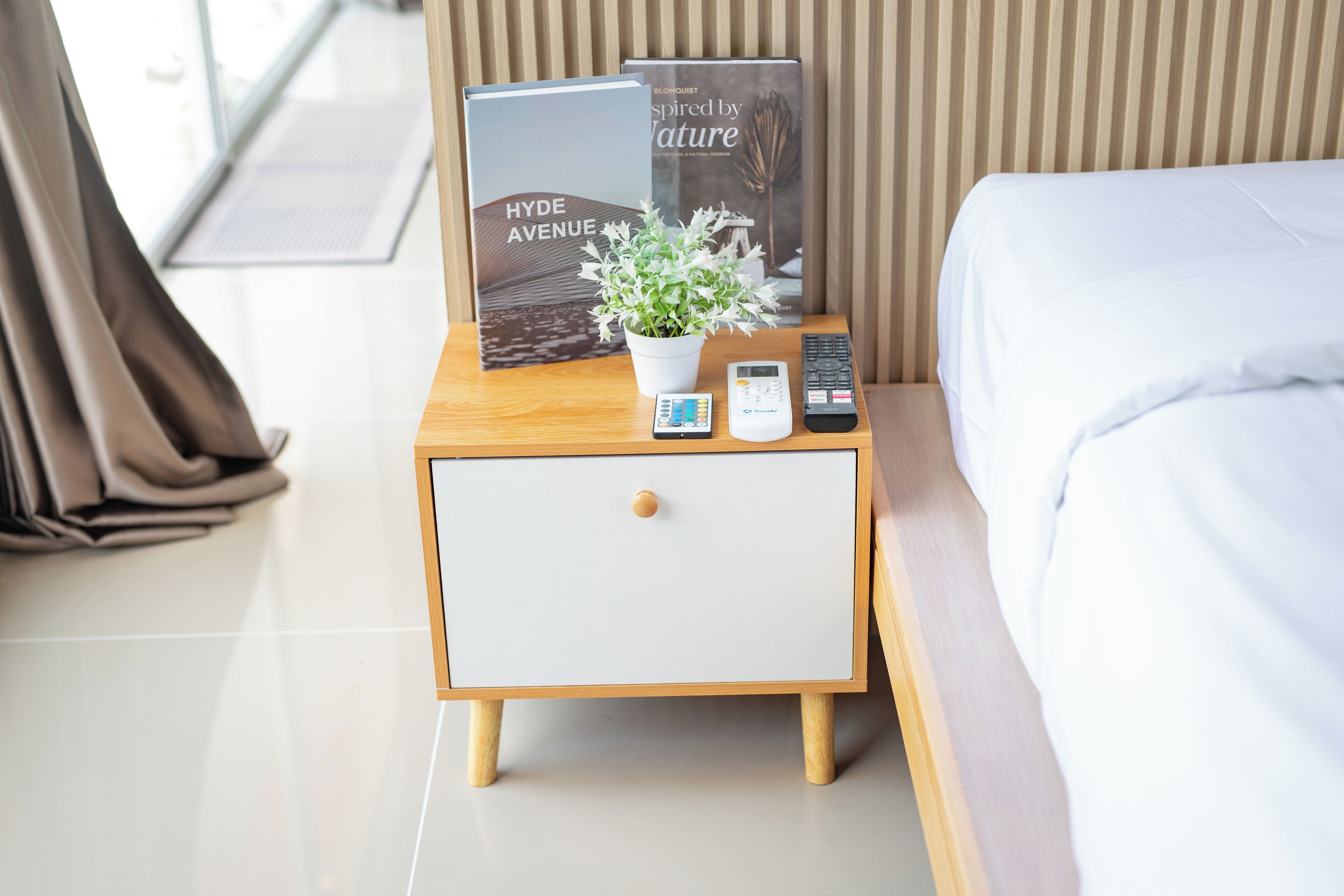 A neatly made hotel room with a bed, a bedside table with a book, a plant, and a remote control.