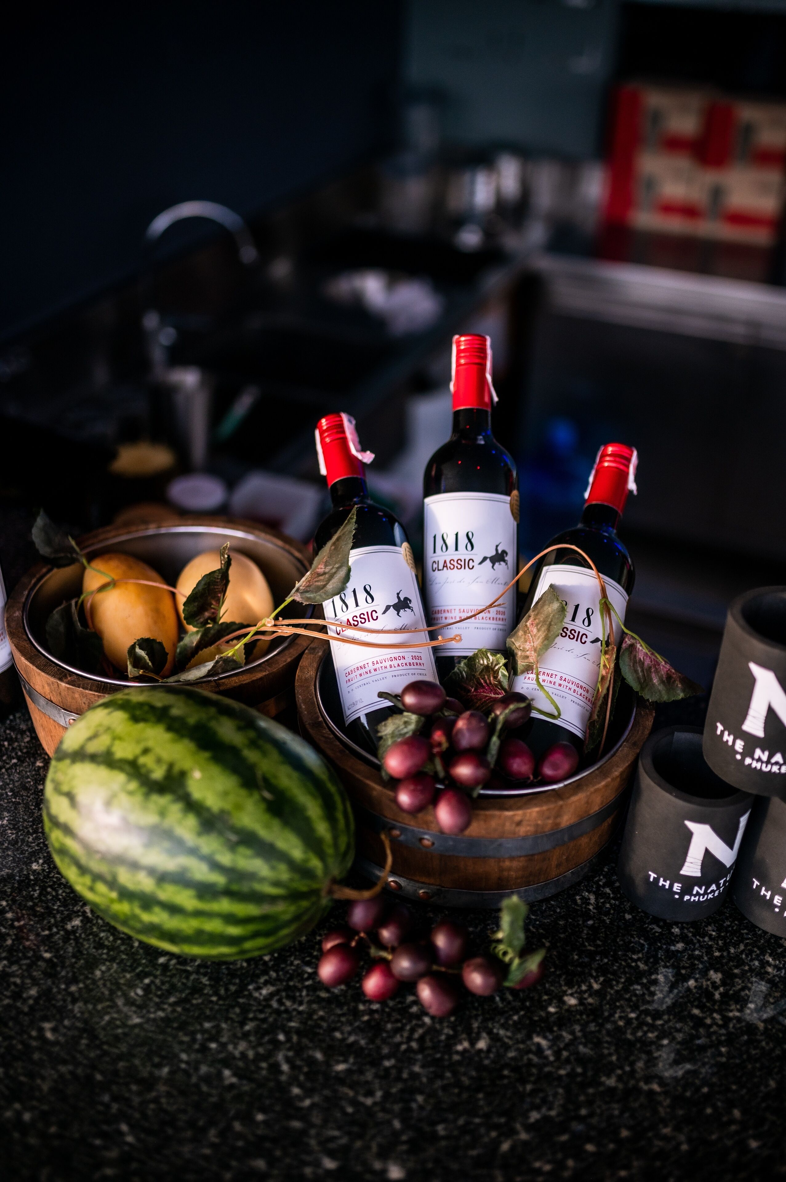 A wooden bowl containing a watermelon, grapes, and a bottle of 1818 Classic wine.