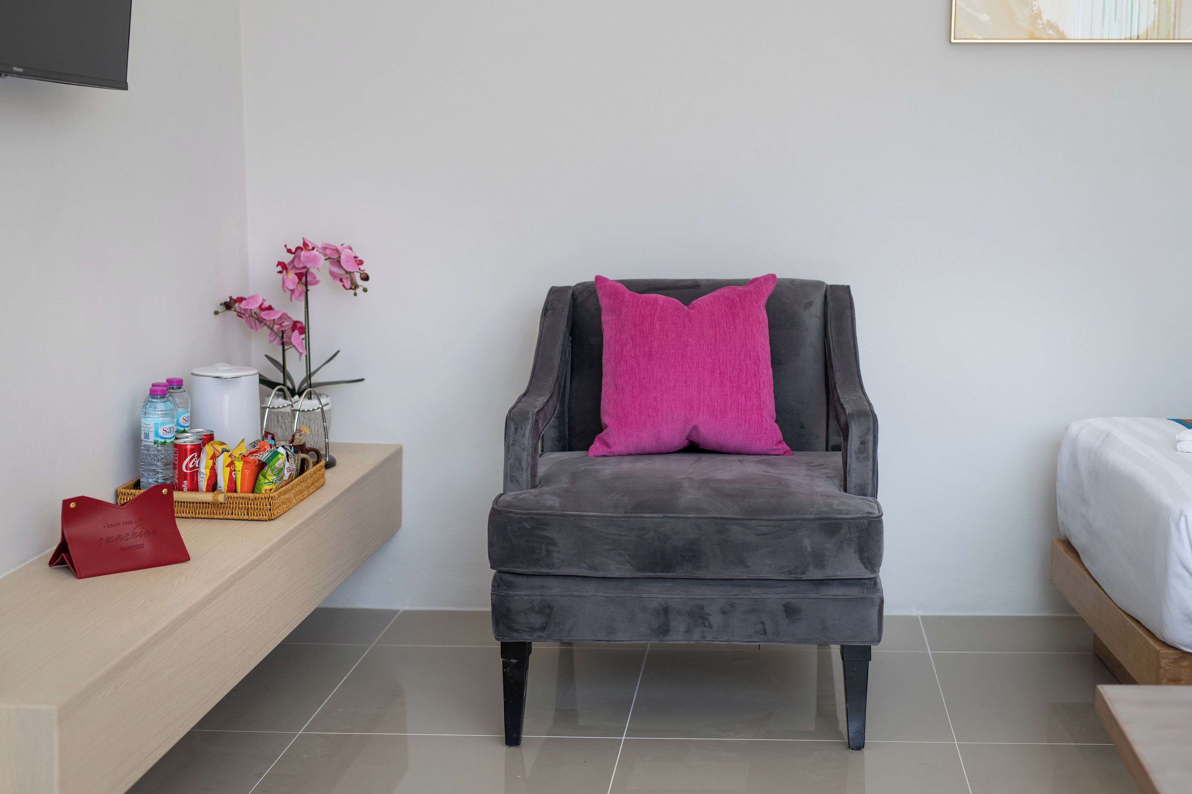 A modern hotel room with a grey armchair, a small side table with snacks and a vase of flowers, and a bed with white bedding.
