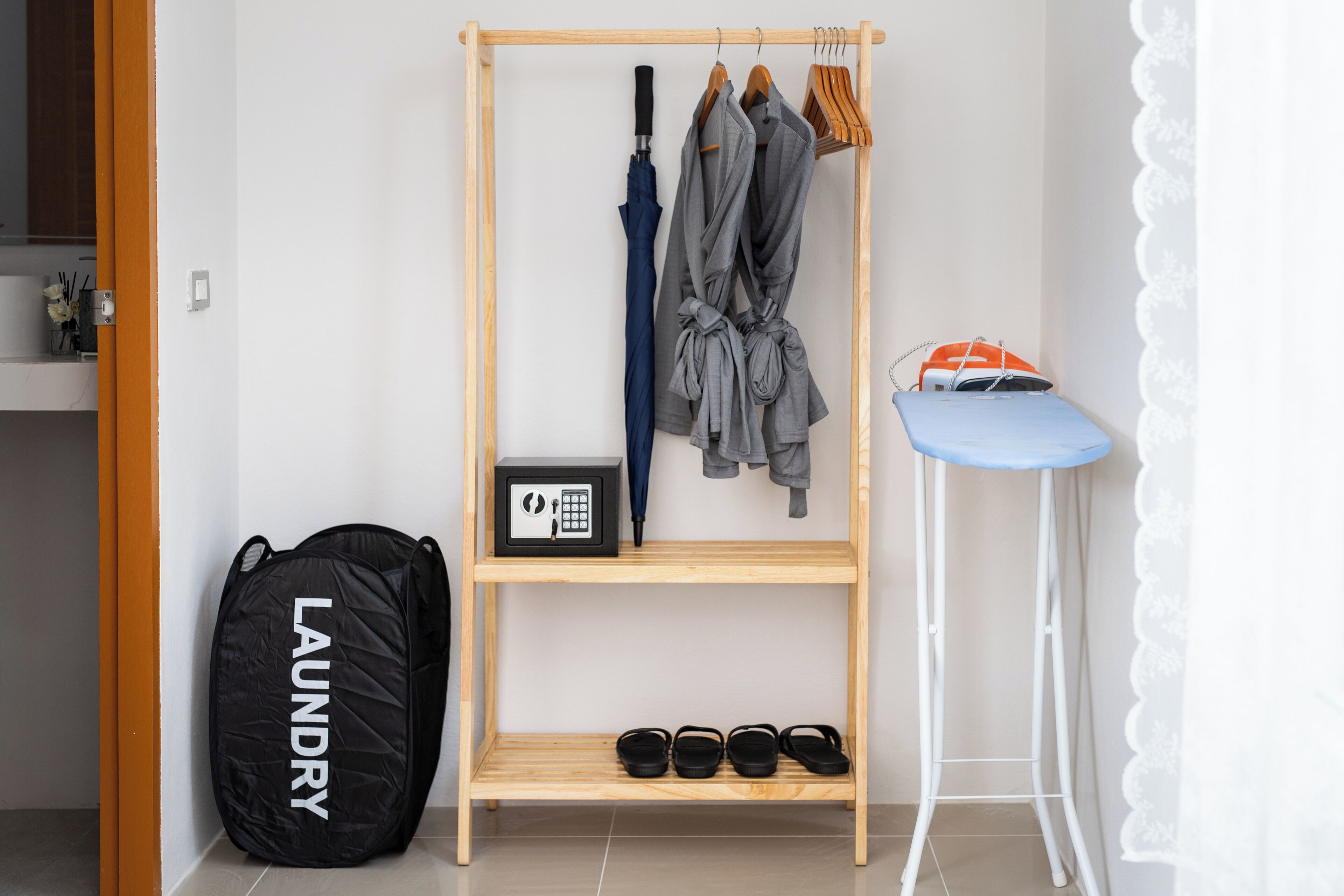 Une armoire en bois contenant des vêtements suspendus, un tabouret bleu et un sac à linge noir.