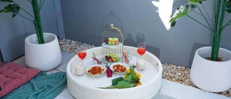 A round table with a white woven tablecloth, surrounded by a variety of food items and drinks, set against a backdrop of a pool and potted plants.