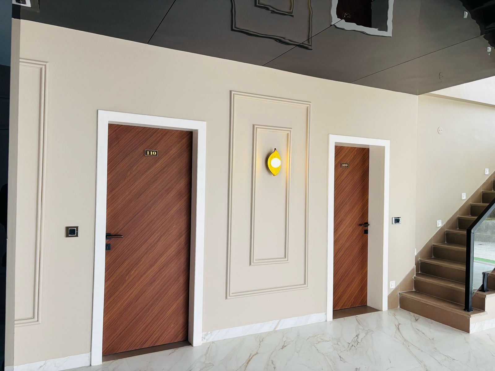 Un pasillo con tres puertas, cada una con una placa de número. Las puertas tienen un acabado de madera, y hay un aplique sobre la puerta central. Las paredes están pintadas en un tono claro y el suelo es de mármol.