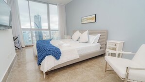 A modern bedroom with a large bed, a white chair, a nightstand, and a view of a cityscape through large windows.