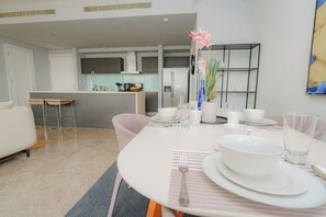 A modern dining area with a white table set for four, a kitchen in the background, and a living area with a sofa and chairs.