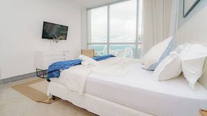 Ein modernes Hotelzimmer mit einem großen Bett, weißer Bettwäsche, einem flachbildfernseher an der Wand und Blick auf die Stadt durch große Fenster.