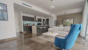 Un salon moderne avec un fauteuil bleu, un canapé blanc et une cuisine en arrière-plan.