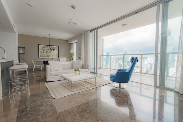 A modern living room with a white sofa, a blue chair, a coffee table, and a city view through large windows. - Exclusive and luxury 1BR with balcony in Yoo Panama (Panamá)