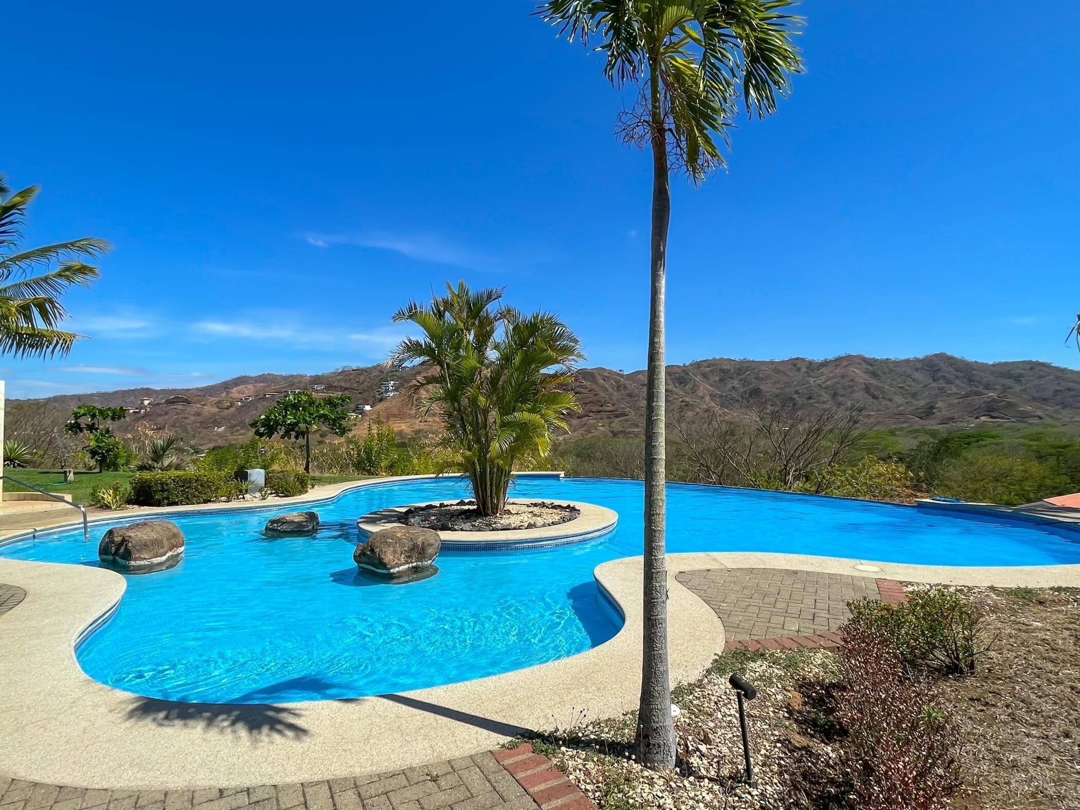 Ein Pool mit gebogener Form, umgeben von Palmen und Felsen, mit Bergen im Hintergrund.