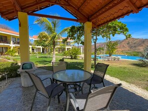 A covered outdoor seating area with a table and chairs, a trash bin, and a view of a pool and mountains in the background. - Colinas Dolce Vita 8017 – 2 Bedroom, 2 Bathroom Condo (Playas del Coco)