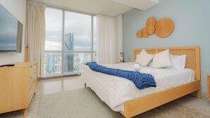 A modern bedroom with a large window, a bed with white bedding and a blue throw, a wooden dresser, and a wall-mounted TV. - Exclusive and luxury 1BR with balcony in Yoo Panama (Panamá)