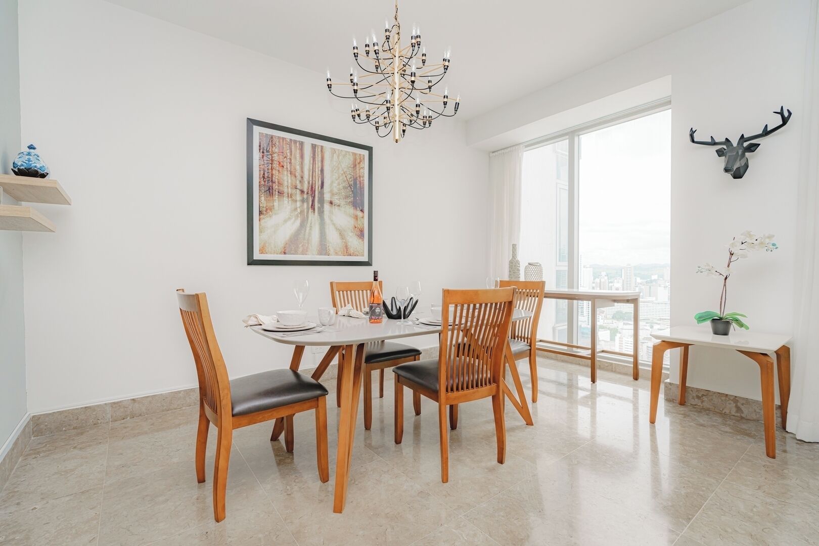 Zona pranzo con un tavolo rotondo, sedie di legno, un lampadario, un quadro incorniciato, vista sulla città attraverso una finestra e una testa di cervo appesa al muro.