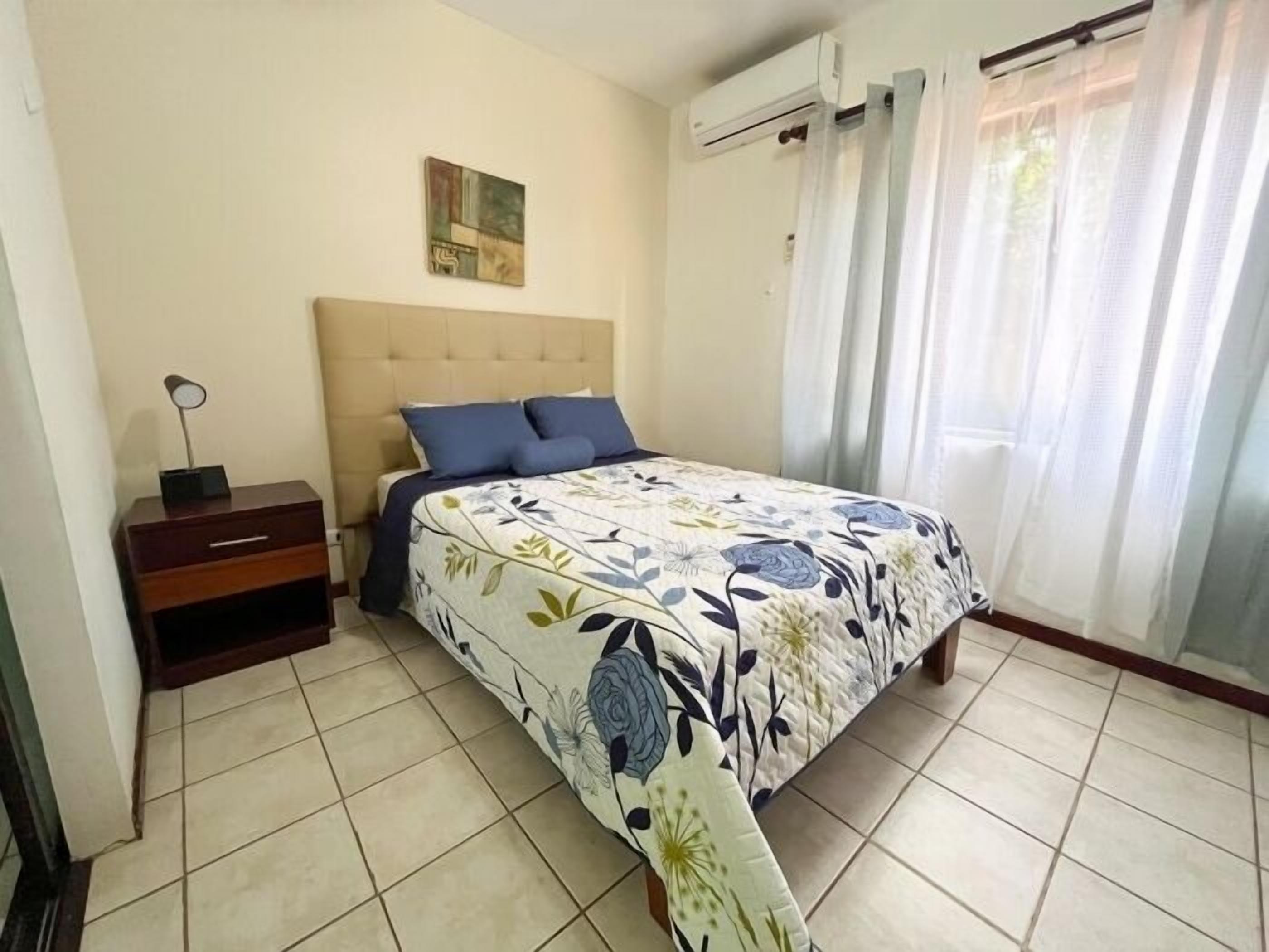 Un dormitorio con cama, mesita de noche, aire acondicionado de pared y ventana con cortinas.