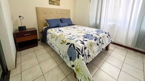 A bedroom with a bed, a nightstand, a wall-mounted air conditioner, and a window with curtains.