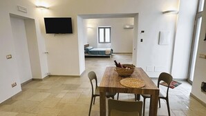 Uma sala de estar ampla com uma mesa de jantar de madeira, quatro cadeiras, uma televisão na parede e uma janela com vista para o exterior.