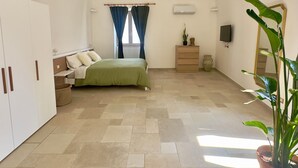 Um quarto amplo com teto alto, uma cama com colcha verde, guarda-roupa, cômoda, uma planta em vaso e um espelho.