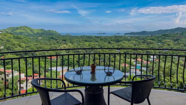 A balcony with a round table and two chairs, overlooking a lush green landscape with distant hills and a clear sky. - Colinas Turquesa L4C Spectacular Ocean-View Vacation Rental (Playas del Coco)