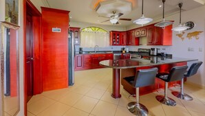 A modern kitchen with red cabinets, a central island with bar stools, and a ceiling fan. - Colinas Turquesa L4C Spectacular Ocean-View Vacation Rental (Playas del Coco)