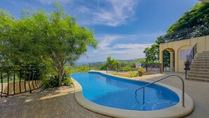 A swimming pool with a curved edge, surrounded by a wooden deck and a metal railing. There is a staircase leading to a covered area with a view of the landscape. - Colinas Turquesa L4C Spectacular Ocean-View Vacation Rental (Playas del Coco)