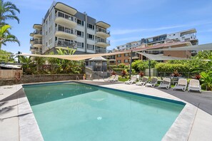 A swimming pool with lounge chairs and a building with multiple balconies in the background. - Cheltenham 4 - Kings Beach Escape With Ocean Views (Caloundra)
