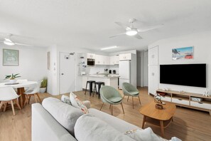 A modern living room with a sofa, chairs, a coffee table, a TV, and a kitchen area with a dining table and bar stools. - Cheltenham 4 - Kings Beach Escape With Ocean Views (Caloundra)