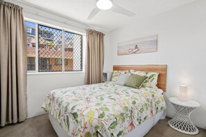 A bedroom with a bed, a window with curtains, a ceiling fan, a small bedside table with a lamp, and a painting on the wall.