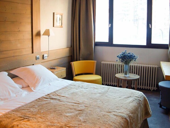 Una habitación de hotel con cama, una silla, una mesita con un florero, un radiador y una ventana con cortinas.