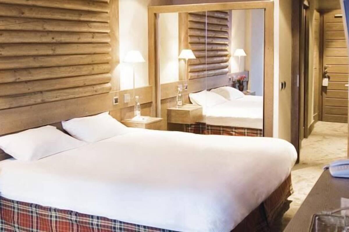 Hotellihuone, jossa on suuri sänky, puinen pääty, yöpöytävalaisimet, peili sekä pieni pöytä, jossa on puhelin ja lasi.