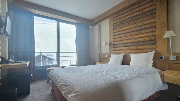 A hotel room with a large bed, wooden headboard, a desk with a telephone, and a view through a window. - Hôtel Le Ski d'Or (Tignes)