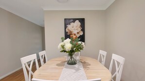 A dining area with a round wooden table, white chairs, a vase with flowers, and a painting on the wall.