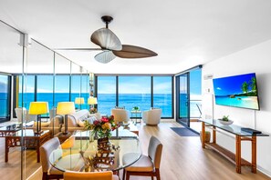 Ein modernes Wohnzimmer mit Glastisch, bequemen Sitzmöglichkeiten, Deckenventilator und großem Fenster mit Meerblick.