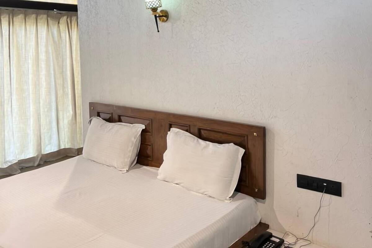 A hotel room with a bed, a bedside table with a phone and remote controls, a wall-mounted light, and a window with curtains.