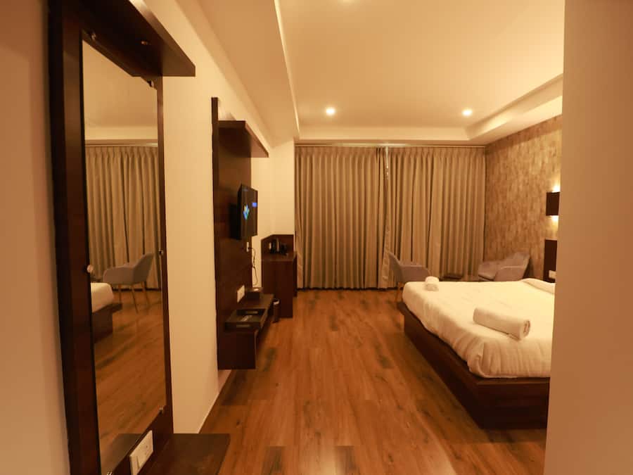 Una habitación de hotel con piso de madera, una cama con sábanas blancas, un televisor sobre un mueble de madera, un escritorio con una silla y un espejo grande.
