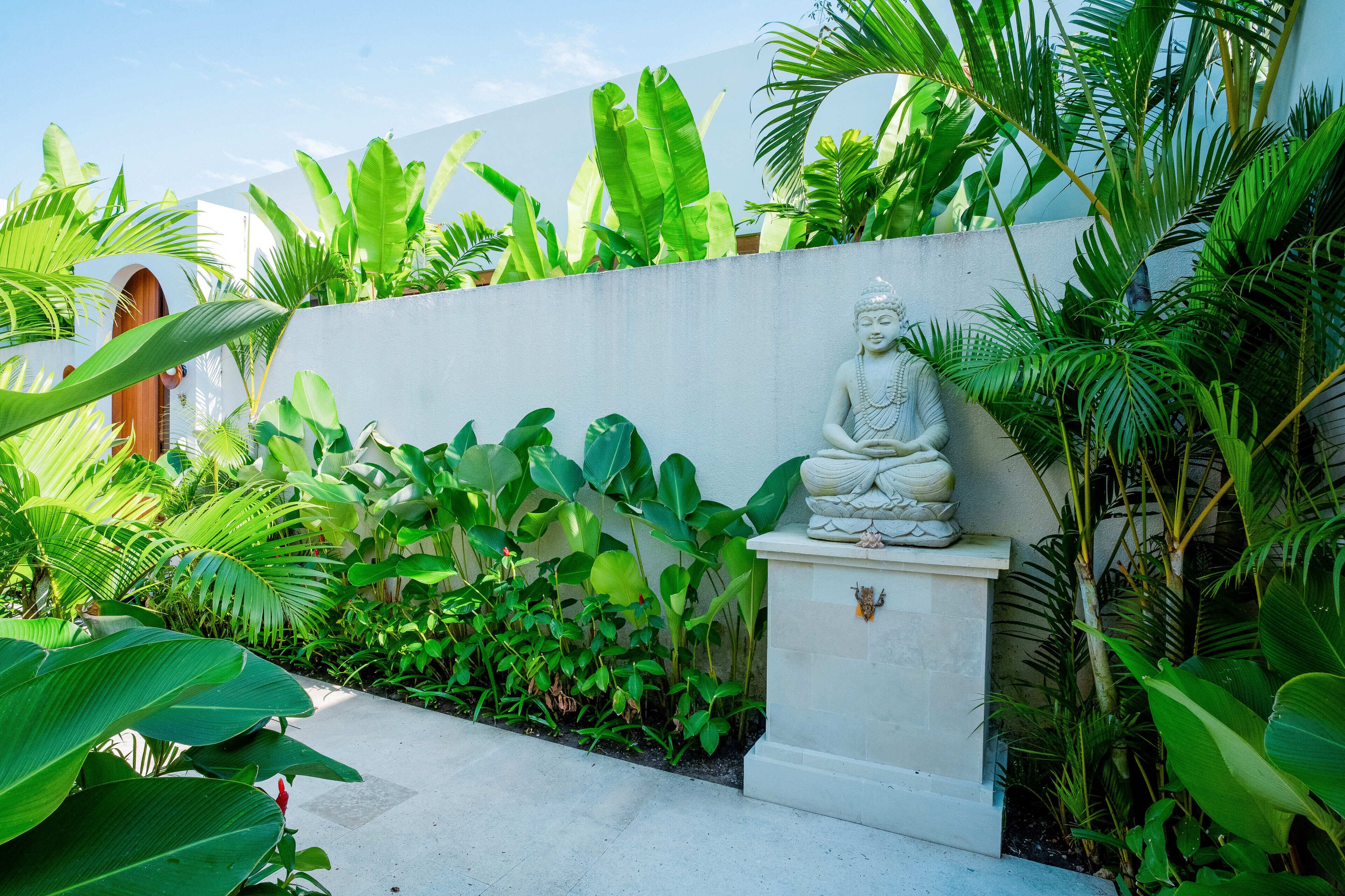 Un espace extérieur paisible, agrémenté d’une statue de Bouddha, d’une végétation luxuriante et d’un mur blanc.