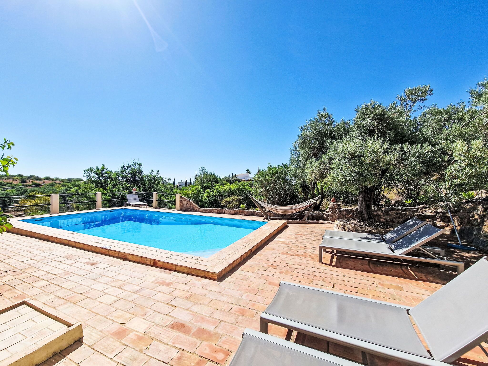 A rectangular swimming pool with a stone border, surrounded by a brick patio. There are lounge chairs and a hammock.
