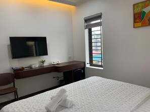 A hotel room with a bed, a TV mounted on the wall, a desk with a computer, and a window with a view of buildings. - Tam Coc TK Hotel  (Nam Hoa Lu)
