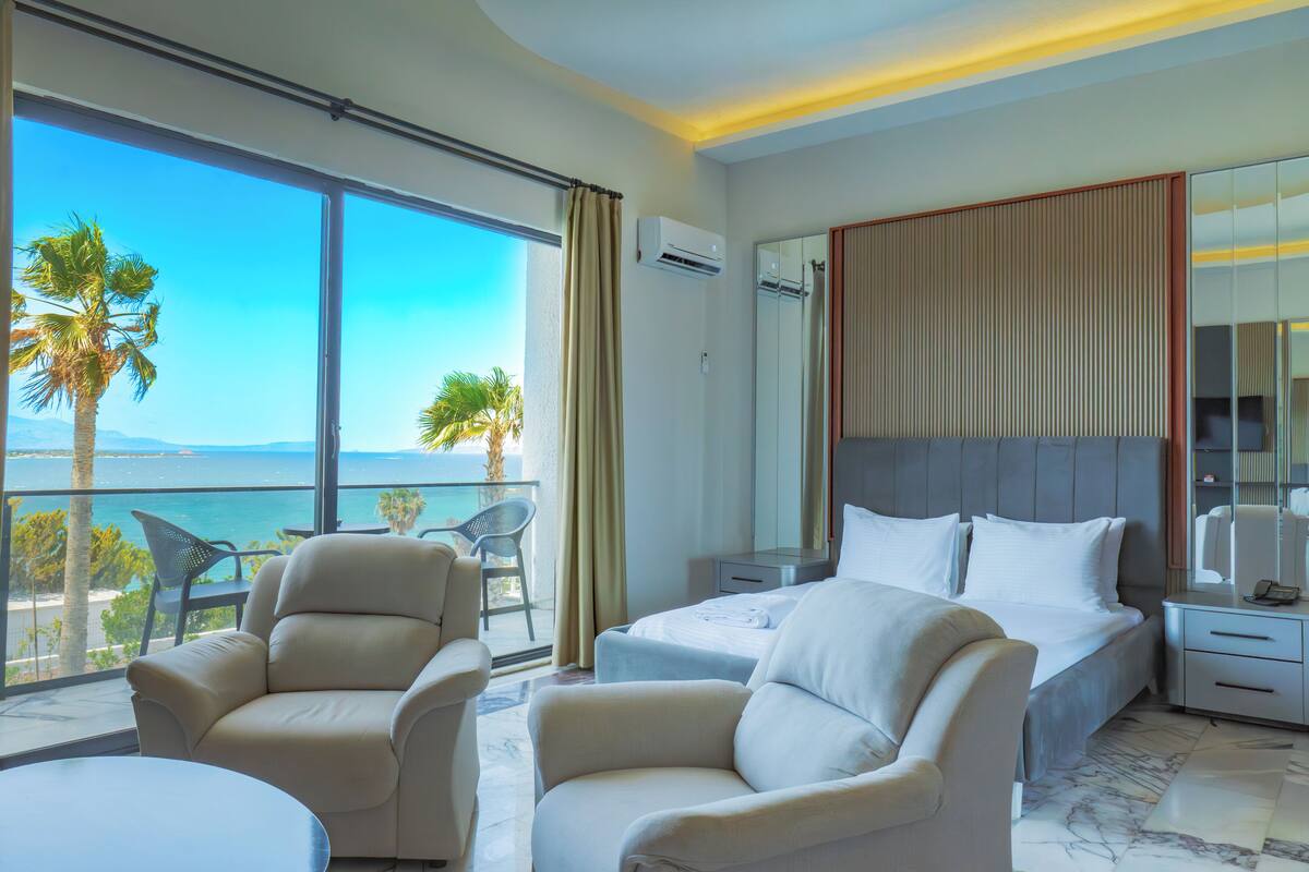 A hotel room with a large balcony, a bed, two armchairs, a nightstand, and a mirror.