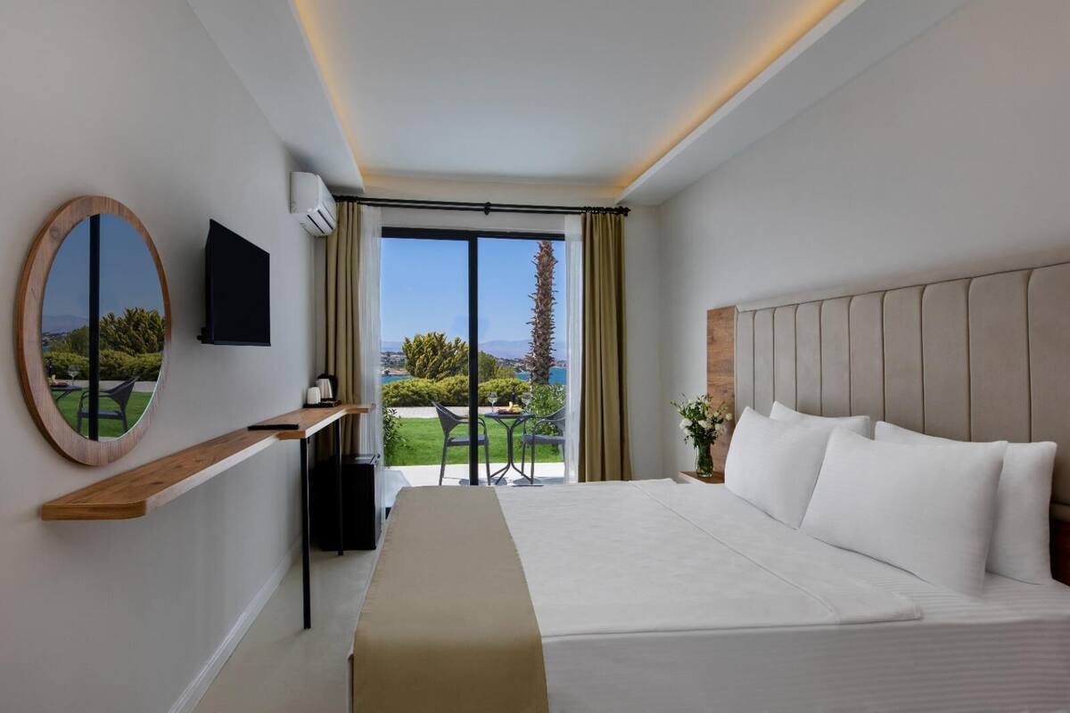 A modern hotel room with a large bed, a TV mounted on the wall, a desk with a chair, and a balcony with a view of the landscape.