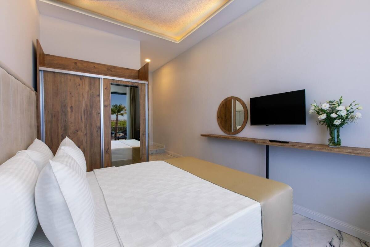 A modern hotel room with a bed, a TV, a mirror, and a wooden shelf with a vase of flowers.