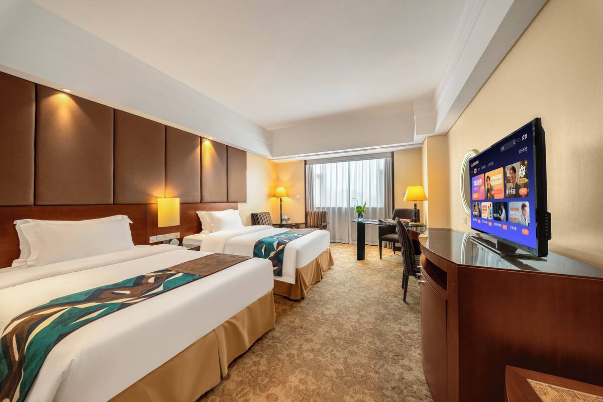 A hotel room with two beds, a desk, a television, and a window with curtains.