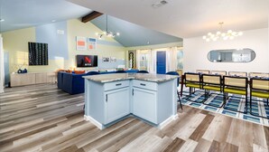 A modern kitchen with a central island, a dining area with chairs, and a living room with a sofa and TV. - The Sunburst: Sleeps 18! Basketball & Heated Pool (San Antonio)