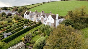 A large, white, two-story house with a well-maintained garden and a green field in the background.
