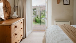 A bedroom with a wooden dresser, a white bed, a balcony with a view of the countryside, and a framed botanical illustration on the wall.