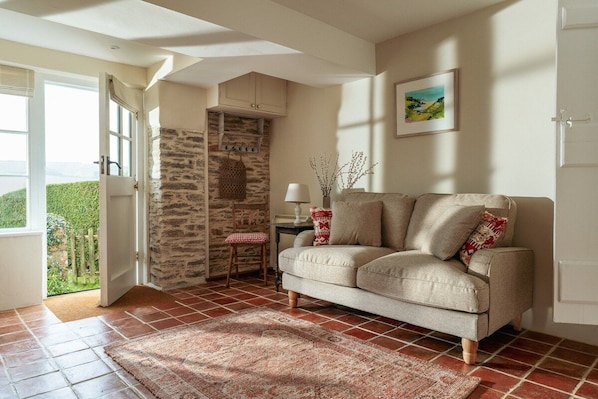 A living room with a sofa, a patterned rug, a stone wall, a window, and a door leading outside.