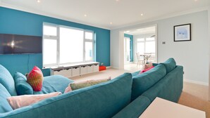 A modern living room with teal sofas, a flat-screen TV mounted on the wall, and a large window providing natural light. - Princes Esplanade (Gurnard)