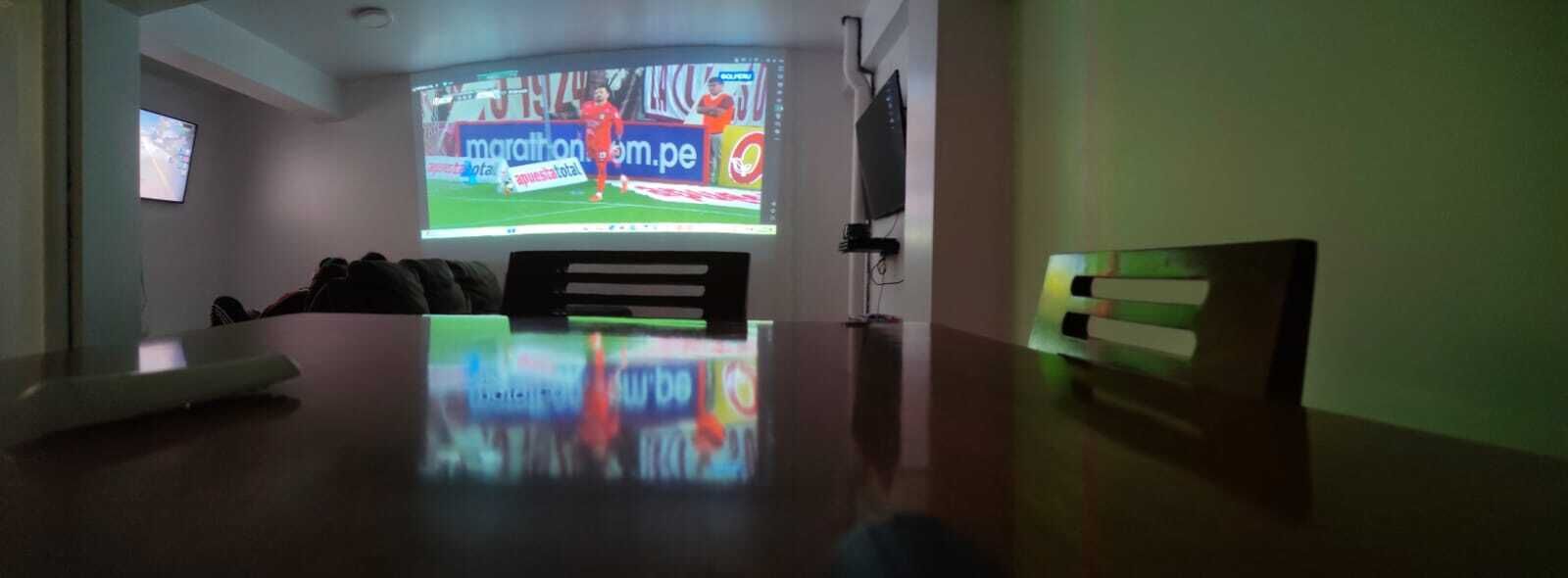 A room with a large screen displaying a sports event, a flat-screen TV, and a wooden table.