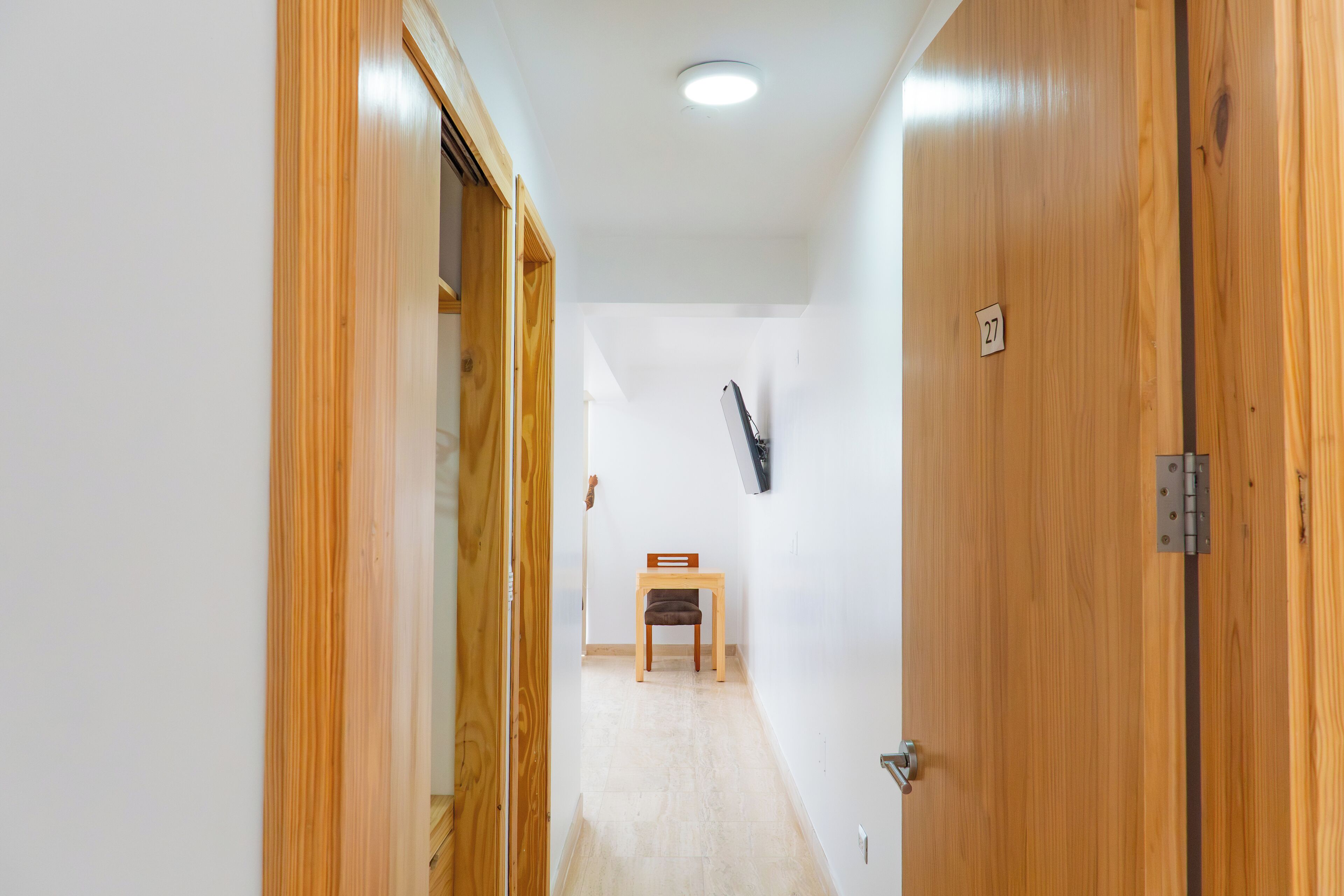 A narrow hallway with wooden doors, a small table with a chair, and a number sign on the wall.