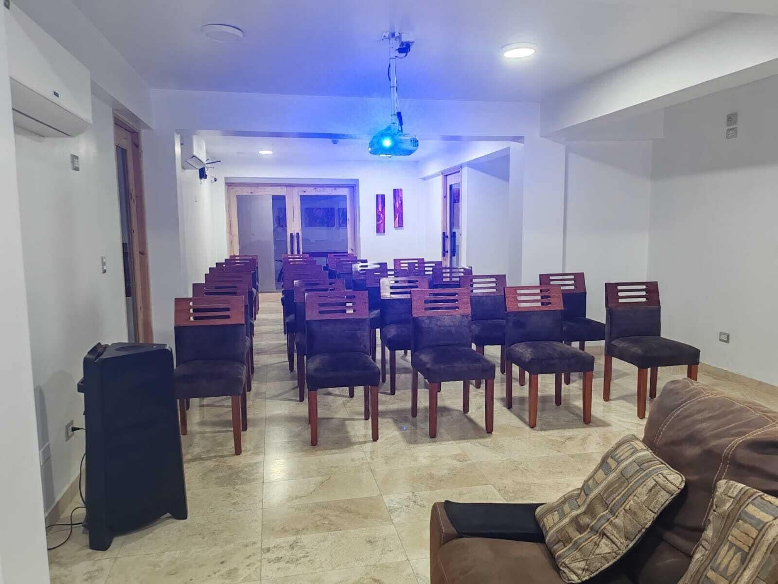 A room with a brown sofa, a dining area with chairs, a ceiling projector, and a tiled floor.
