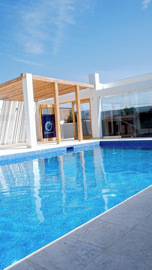 A modern outdoor swimming pool with a wooden pergola and glass doors, surrounded by a white building. -  Hotel Boutique La Angostura II (Ica)