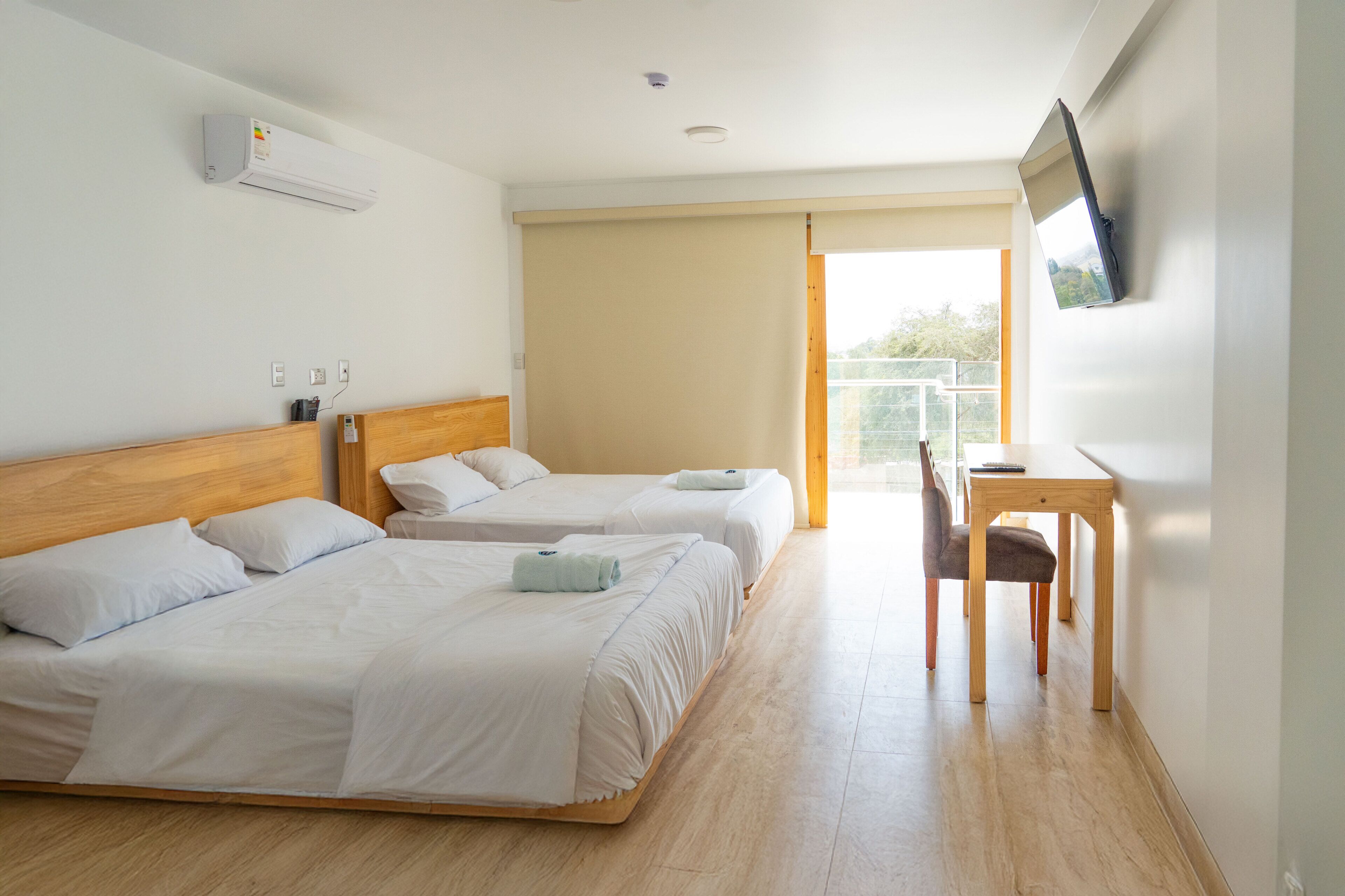 A hotel room with two beds, a desk, a chair, an air conditioning unit, and a television mounted on the wall.
