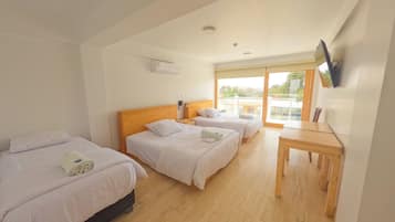 A hotel room with three beds, a wooden table, a TV mounted on the wall, and a window with a view of the outside.